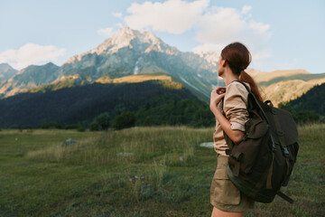 Naklejka premium Woman with backpack standing in a grassy mountain meadow, exploring outdoors, warm light, distant peaks, casual clothing, serene mood, natural colors, and sense of adventure.