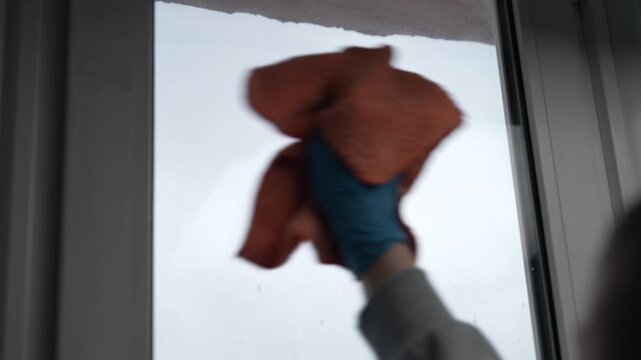 Gloved hand cleaning dirty glass window pane with orange microfiber cloth. Housekeeping professional performing routine chores for spotless and transparent home surface. Shooting in slow motion.