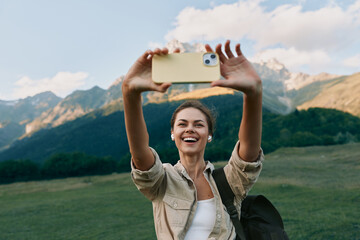 Naklejka premium Young woman lifestyle traveler takes selfie with smartphone on mountain meadow background, smiling happy tourist enjoys nature and outdoor adventure in sunny day.