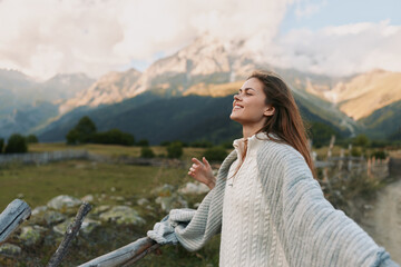 Naklejka premium A woman stands outdoors on a sunny mountainside, arms outstretched, smiling, wearing a cozy sweater. Bright landscape highlights freedom, happiness, and a carefree lifestyle.