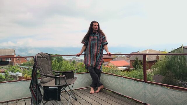 Eco-Conscious Millennial Traveler Wearing Tribal Serape and Harem Slacks Standing on Veranda Leaning Against Railing Observing Scenic Mountain Bay and Cloudy Sky View Authentic Lifestyle Wide Shot