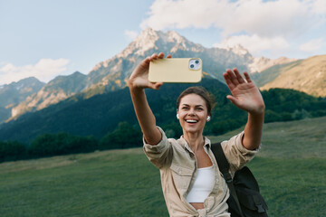 Naklejka premium Cheerful woman outdoors taking a selfie in a mountain meadow, bright smile, casual outfit, sunny day, sense of adventure, natural colors, scenic backdrop amid lush greens