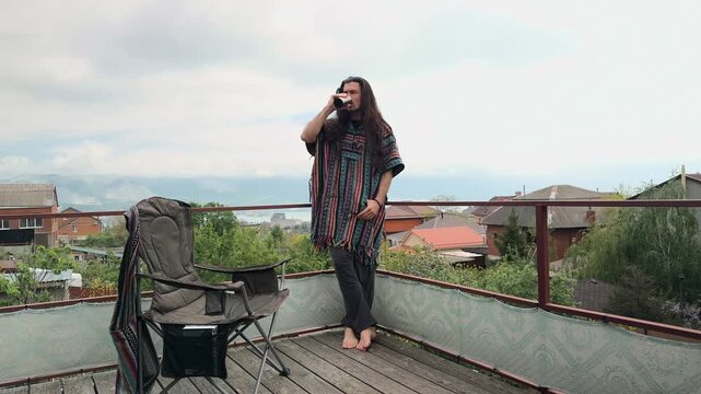 Free-Spirited Man with Long Messy Hair Wearing Folk Patterned Baja Poncho and Grey Dhoti Pants Standing on Terrace Taking a Sip from Silver Thermos Tumbler Enjoying Scenic Coastal Mountain Harbor