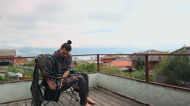 Pensive Man with Man Bun Wearing Ethnic Poncho Putting Book in Camping Chair Side Pocket and Leaning Forward in Thinker Pose on Terrace with Scenic Sea and Mountain View Looking Very Thoughtful