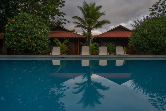 View of a swimming pool reflecting wooden bungalows with red roofs, white sunbeds, and a tall palm tree under a cloudy evening sky.