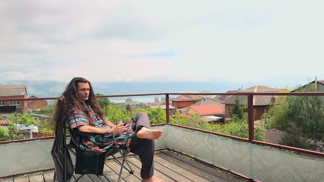 Wild Spirit Bohemian Man with Long Loose Hair Blowing in Wind Sitting on Terrace Holding Silver Thermos Mug Enjoying Spring Day View of Sea and Mountains Wearing Ethnic Patterned Poncho and Pants