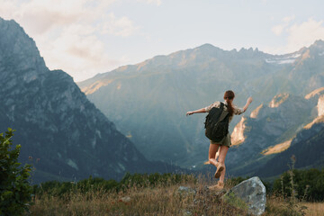 Naklejka premium A young woman with a backpack strides through a grassy meadow toward dramatic mountains, radiating adventure, freedom, and focus on the trail ahead.