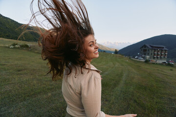 Naklejka premium Happy woman outdoors turning with hair flying, smiling joyfully, wearing casual clothes, green meadow, mountain landscape, clear sky background