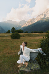 Naklejka premium A woman sits on a rock in a white knit sweater and boots, stretching her arms, gazing toward distant mountains, a calm outdoor lifestyle moment in nature.