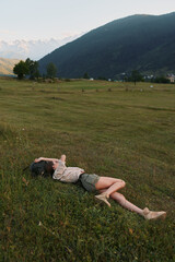 Naklejka premium Lying woman in a sunlit meadow, relaxed pose in a wide field with distant mountains, celebrating outdoor lifestyle, natural colors and peaceful countryside under blue sky warm breeze vivid greens.