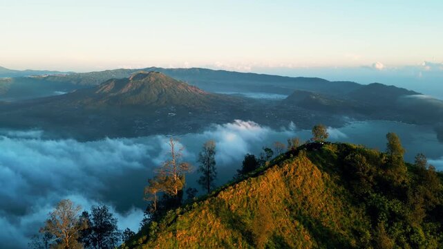 Bali volcano mountain ridge with crater lake above clouds in golden light, low fog drifting through valley with slow descending aerial movement over volcanic terrain, aerial footage