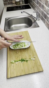 Using a knife to transfer chopped green onions from a board to a white oblong plate. Aesthetic greenery preparation process.