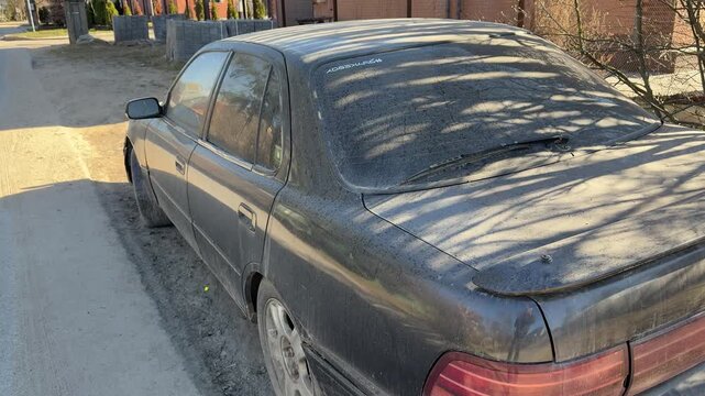 Dusty black sedan abandoned by roadside, frontend collision damage with cracked bumper, tinted windows covered in grime