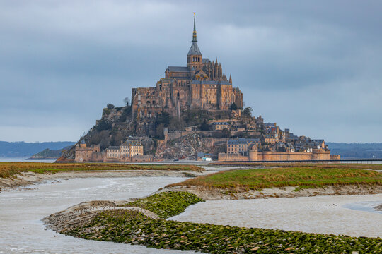 View of Mont Saint-Michel abbey rising above the tidal flats and muddy channels under a cloudy sky Mont Saint-Michel, Normandy, France.