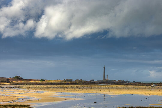 View of Ile Vierge lighthouse standing tall on a rocky horizon under a dramatic cloudy sky with a sandy beach and tidal pools in the foreground in Plouguerneau, Bretagne, France.