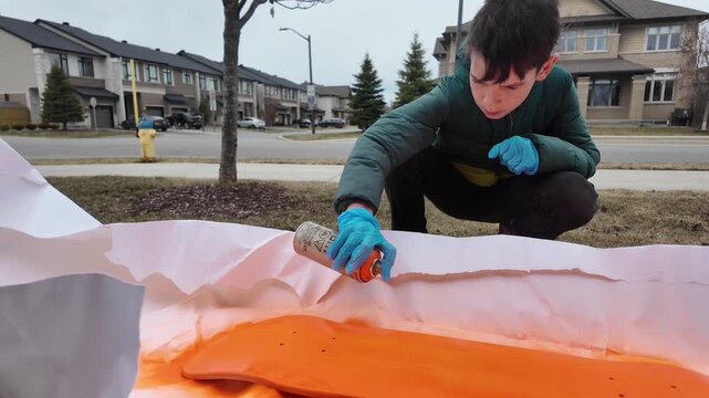 Kid spray paints skateboard deck on front lawn - wide shot of him focusing and covering full deck