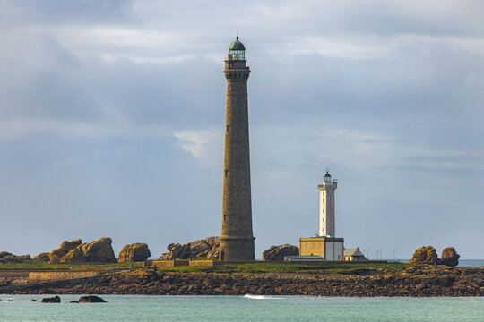 View of the Ile Vierge lighthouse with its tall stone tower and shorter white companion standing on a rocky island under a cloudy sky in Plouguerneau, Bretagne, France.