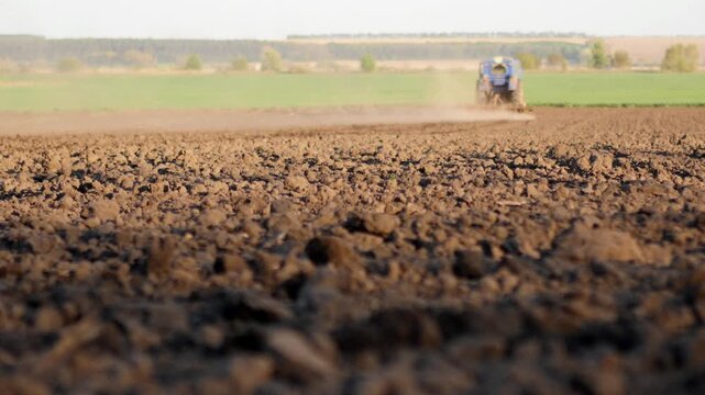 Close-up of brown soil clods in the foreground with a blue tractor working in the distance. Shallow depth of field creates a cinematic look for agricultural and environmental themes