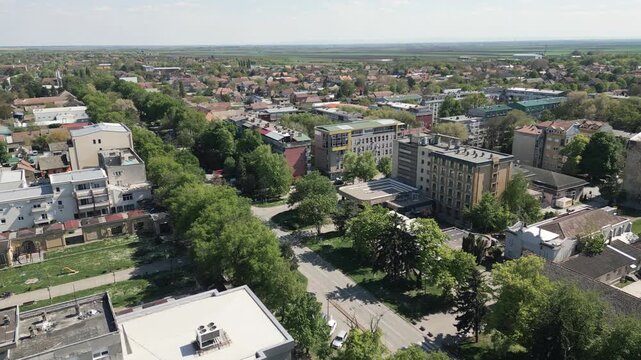 High altitude aerial shot flying forward over the center of Vrbas, Serbia. Wide panorama features a prominent tree lined avenue cutting diagonally through the cityscape on a bright sunny day.