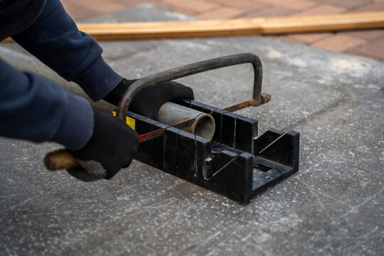 Worker in gloves cutting a metal pipe with a hacksaw in a miter box on a concrete work surface.