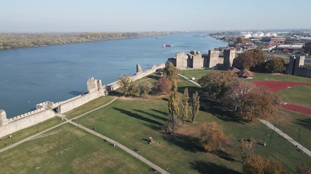 Sunny aerial shot flying forward over the green park inside Smederevo Fortress, Serbia. View of well preserved medieval stone walls and the Danube river under a clear blue sky.