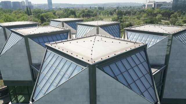 Low altitude aerial close-up of a brutalist building roof in New Belgrade. Focus on sharp edges, triangular concrete shapes, and geometric patterns under bright sunlight. Socialist-era architecture.