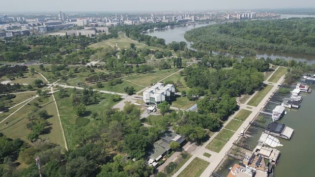 High altitude aerial shot flying forward over the green landscape of Usce Park in New Belgrade. Panorama of the river confluence and distant city buildings on a bright sunny day in Serbia.