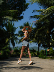 Naklejka premium young woman with curly hair wearing sunglasses and denim shorts jumping on a sunny tropical path surrounded by palm trees and clear blue sky