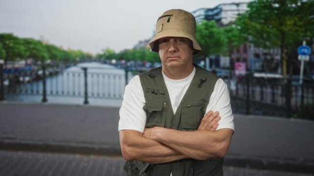 Man with folded arms wearing fishing vest, bucket hat and white shirt on canal bridge with railing and water; quiet stoicism.