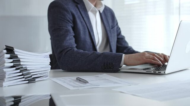 Businessman typing on laptop amid stacked documents and reports, focused on financial data and paperwork in a modern office, conveying productivity, management, and analysis. Audit and taxes