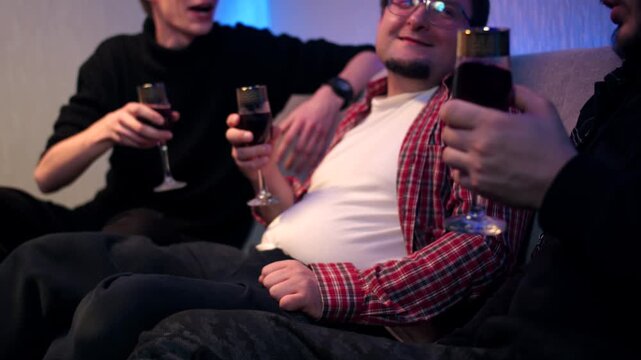 Group of happy friends laughter and toasting in domestic room with neon light. Joyful gathering of caucasian young men in casual cloth on sofa and clink drinks. Drinking red wine in glasses at home