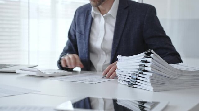 Businessman sorting through stacks of documents on a white desk beside a tall pile of bound papers, symbolizing meticulous financial record keeping and bureaucracy. Audit and taxes