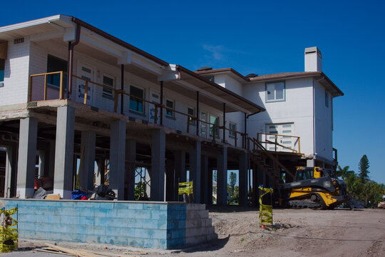 Elevated coastal home raised on concrete stilts undergoing renovation with heavy machinery, exposed foundation, temporary barriers and construction materials at a sunny seaside property preparing for 