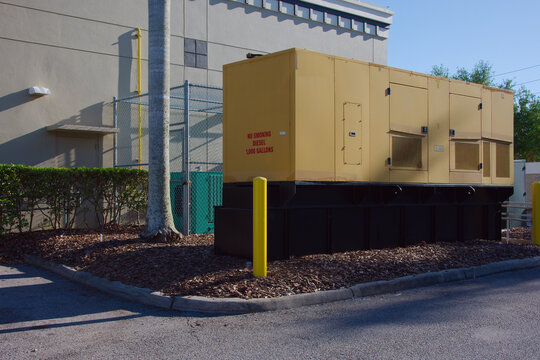 Large yellow diesel backup generator sits on a raised platform beside a commercial building, surrounded by mulch, safety bollards and fencing, illustrating infrastructure, emergency power and industri