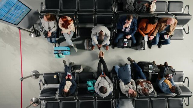 Bored multiethnic travelers in airport terminal waiting for flight status updates on departure board. Passengers annoyed by flight delay, waiting for boarding call, high angle shot