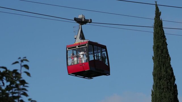red funicular car leading to the top of the old mountain. The tourist car rides above rocky mountains dotted with cypress trees. Cable car cabins amazing sky and clouds. Cableway, green transportation