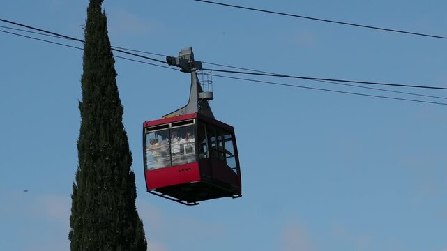 red funicular car leading to the top of the old mountain. The tourist car rides above rocky mountains dotted with cypress trees. Cable car cabins amazing sky and clouds. Cableway, green transportation