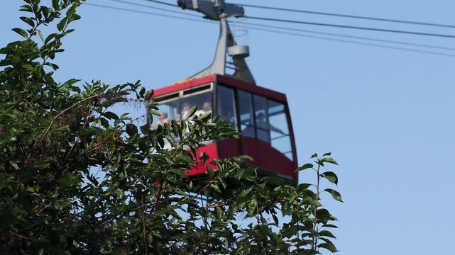 red funicular car leading to the top of the old mountain. The tourist car rides above rocky mountains dotted with cypress trees. Cable car cabins amazing sky and clouds. Cableway, green transportation