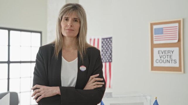 Woman wearing i voted sticker with arms crossed beside a transparent ballot box and american flag in a building; civic pride.
