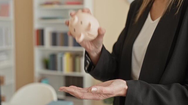 Woman holding pale piggy bank above open palm inside building, wearing business blazer and ring, gesturing offering savings; financial prudence.