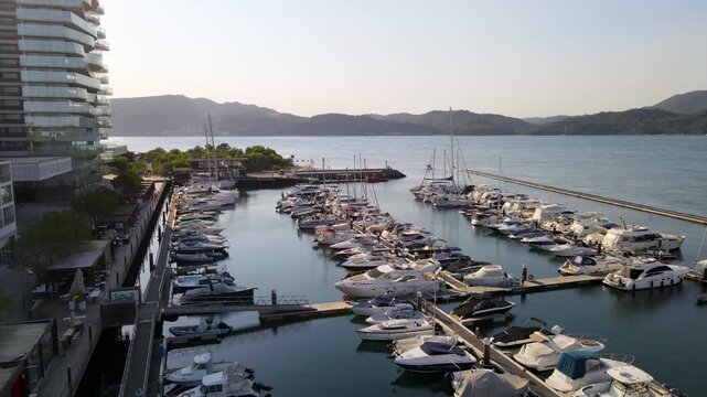 Cinematic drone shot flying at low altitude over parked sailboats and motorboats in Troia Marina. The footage captures the golden hour light over the harbor, modern waterfront architecture, and the tr