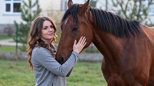 Young woman with long curly hair gently caressing a brown horses face in a rural outdoor setting.
