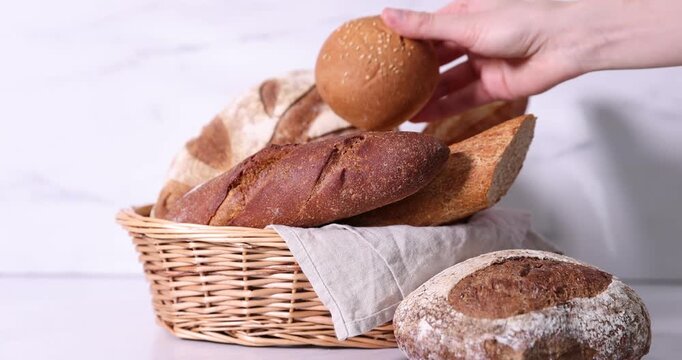 Woman taking fresh bun from wicker basket with different types of bread at white countertop, closeup