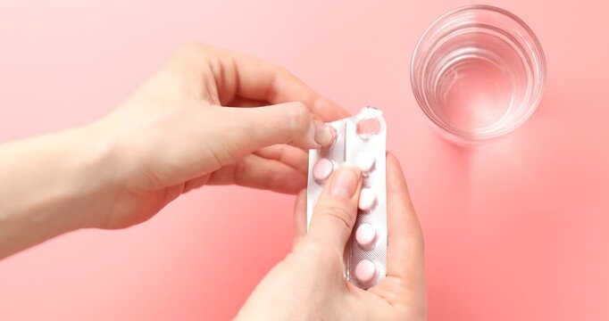 Woman taking pills out from blister and glass of water on pink background, top view