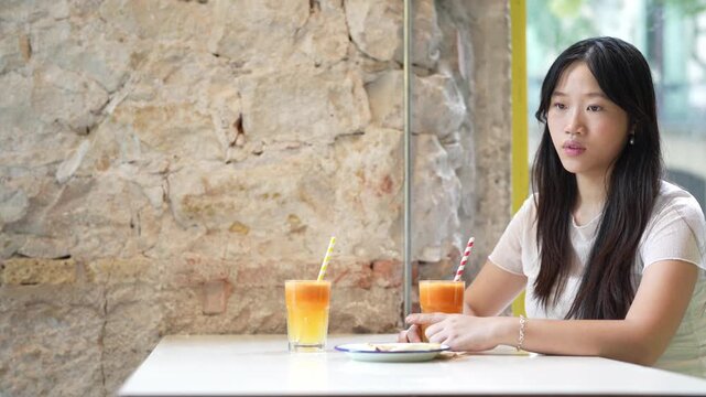 Young woman sits quietly at cafe table. She gazes thoughtfully past the camera. Two orange drinks rest before her. Her expression suggests contemplation or calm boredom