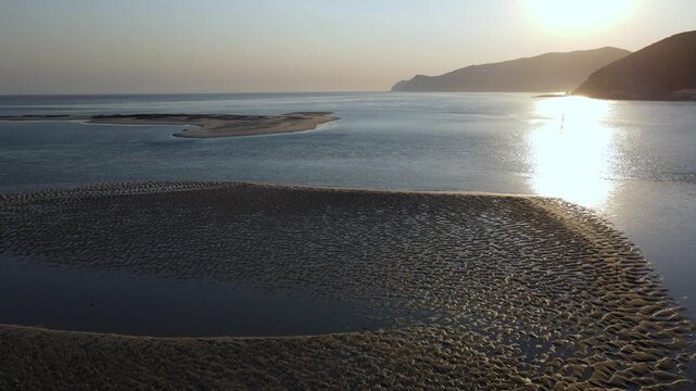 Aerial view of a flock of birds flying up from a sandbank at sunset. Drone flying forward towards Arrabida mountains, Troia, Portugal.