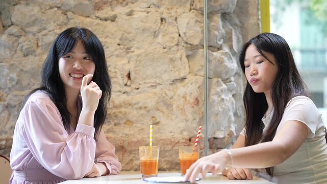 Two women laugh at a rustic cafe table. One smiles broadly, eyes bright with joy. The other sips a drink, eyes half-closed in amusement. Orange juice and straws sit before them
