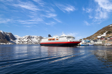 A large Norwegian Cruise ship with red hull docked at Honningsvag port, Norway, with snow-covered mountains and a small Arctic town visible under a bright blue sky © eskystudio