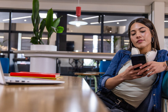 Woman in denim jacket and headphones checking smartphone at communal table near laptop, copy space