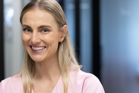 Adult woman smiling, facing camera, wearing light pink blouse and gold hoops in office corridor